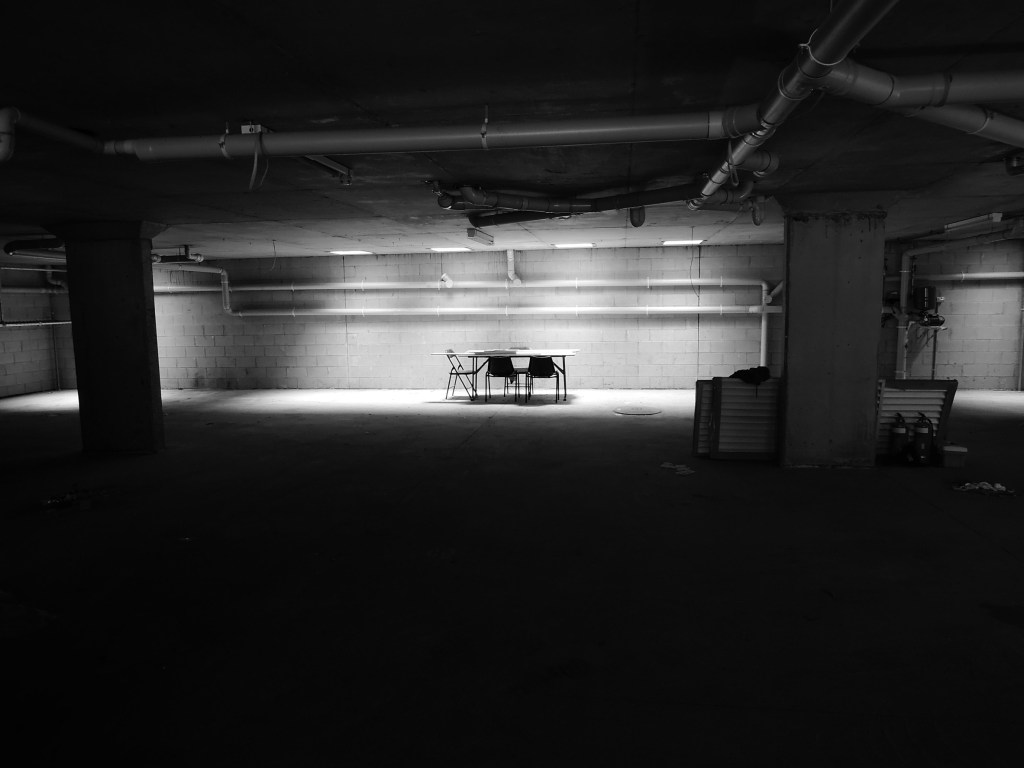 A dark basement with a far-off brightly lit folding table in the center and four folding chairs around it
